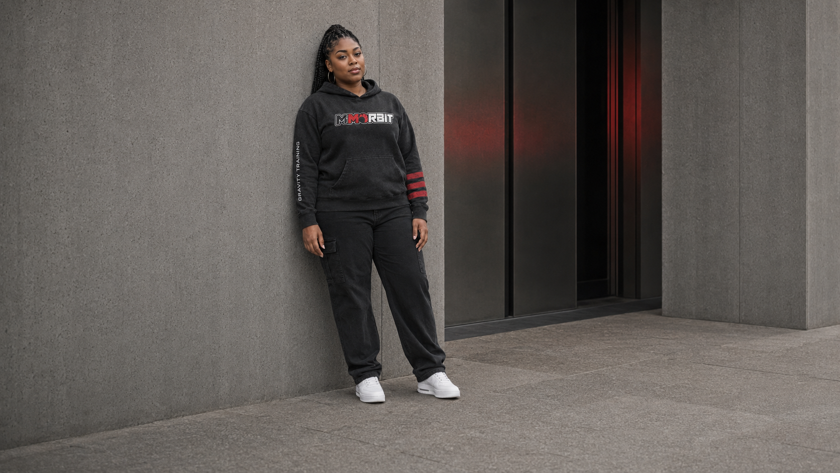Full-body view of one adult woman wearing a naturally fitted MMORBIT Gravity Training hoodie in a relaxed concrete-wall portrait