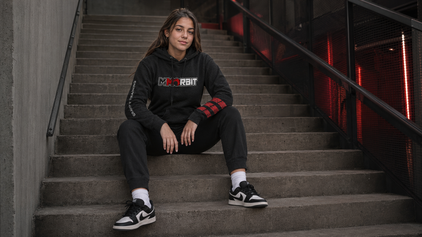 One adult older-teen model seated naturally on industrial stairs in a contoured MMORBIT Gravity Training hoodie